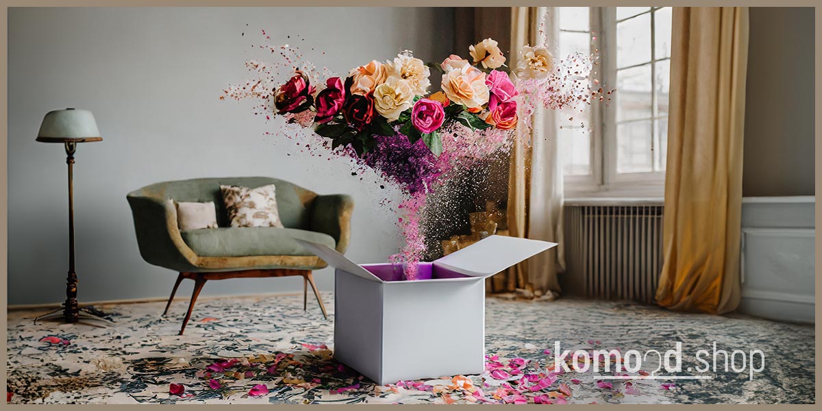 Abbildung eines Geschenkgutscheins mit einem mit Blumen explodierenden Geschenk. Im Hintergrund ist ein helles Wohnzimmer im schlichten Stil zu sehen. Angebot aus dem komood.shop.